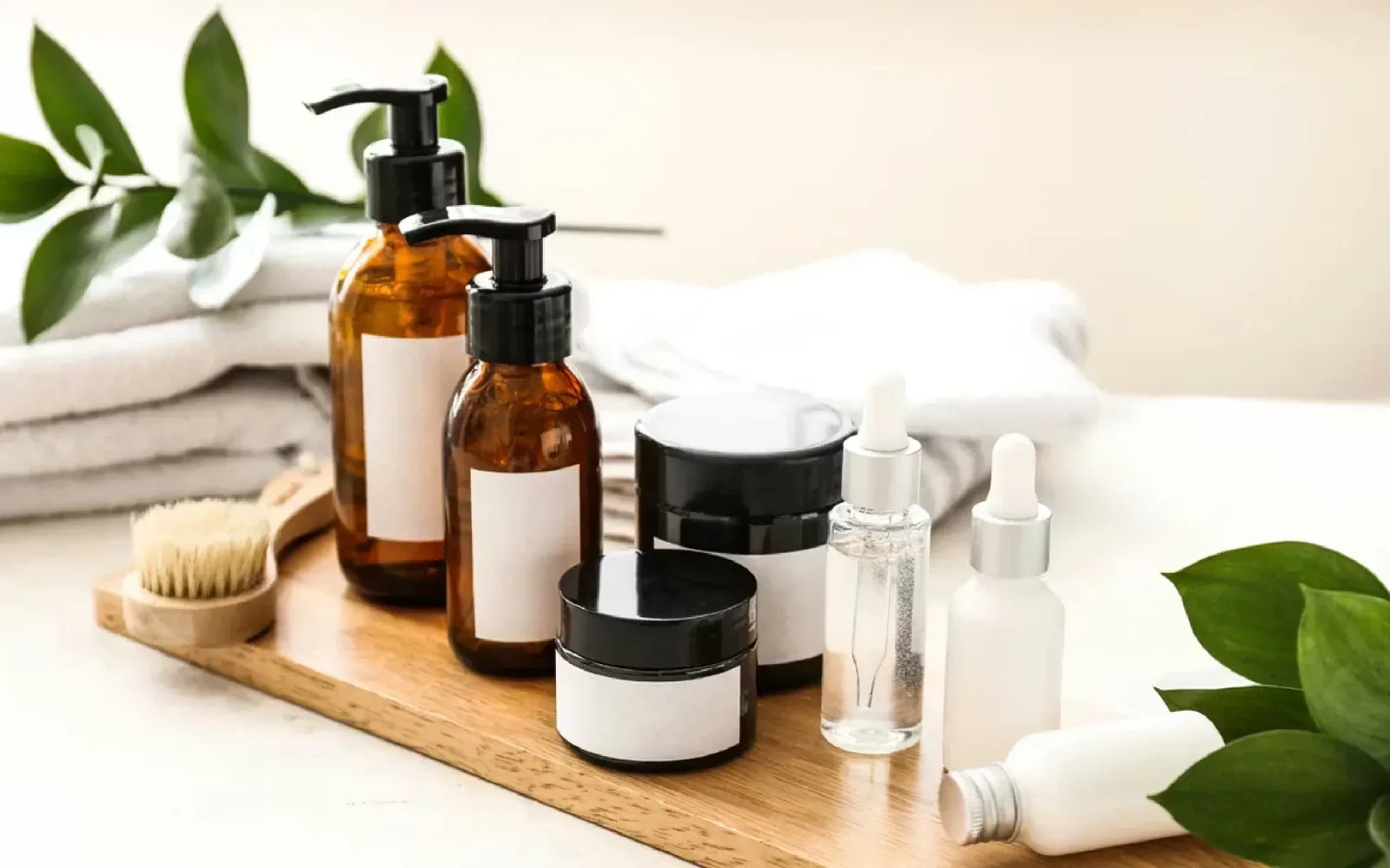 Productos de cuidado facial en frascos de vidrio ámbar y blanco sobre una bandeja de madera, junto a hojas verdes.