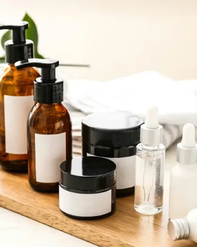 Productos de cuidado facial en frascos de vidrio ámbar y blanco sobre una bandeja de madera, junto a hojas verdes.