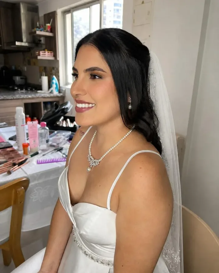 Novia sonriente con maquillaje luminoso en tonos suaves y labios rosados, con velo, collar de perlas y vestido blanco.