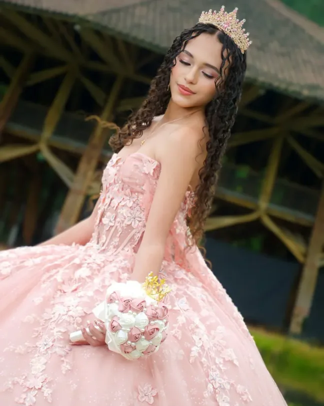 Quinceañera con maquillaje en tonos rosados, corona dorada, cabello largo rizado, vestido rosa bordado con flores y ramo en la mano.