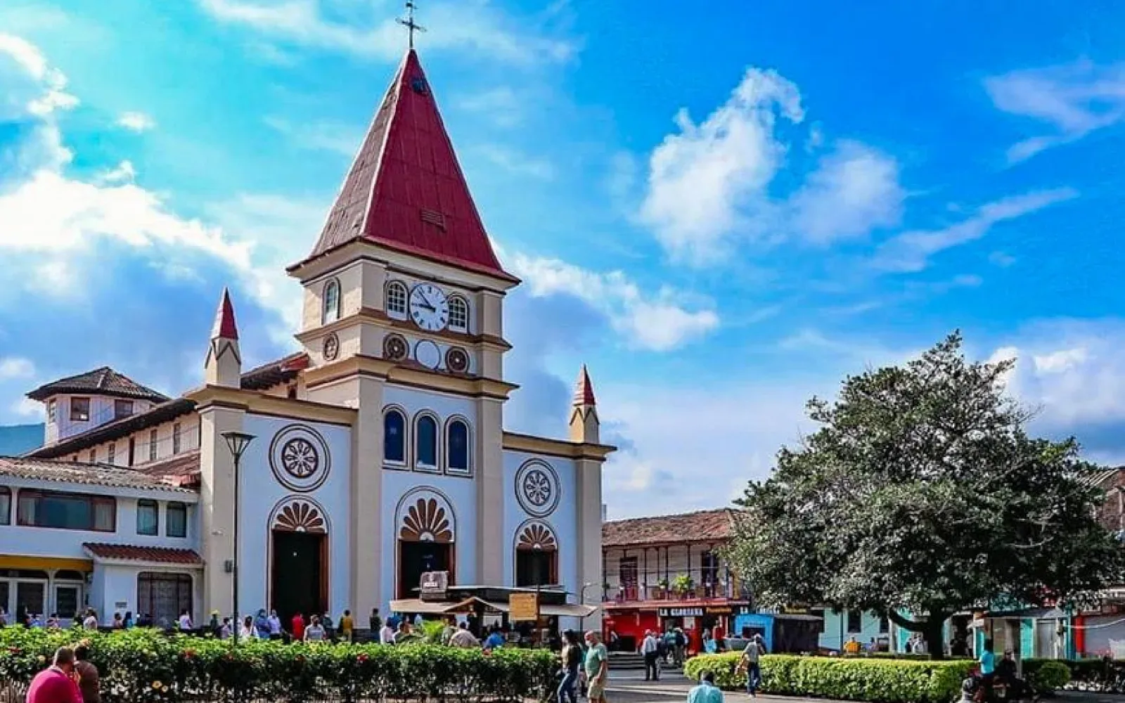 Iglesia de fachada blanca con torre de reloj y techo rojo frente a una plaza arborizada.