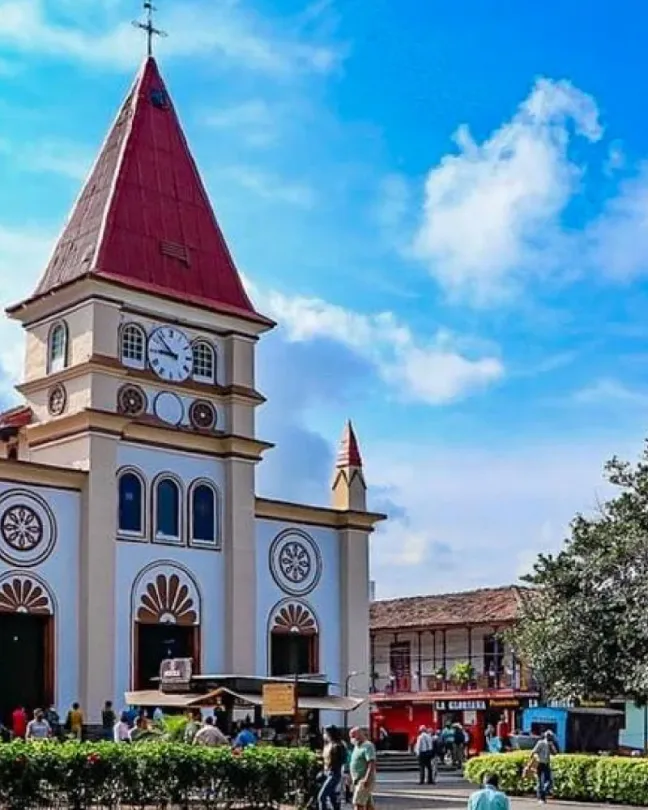 Iglesia de fachada blanca con torre de reloj y techo rojo frente a una plaza arborizada.