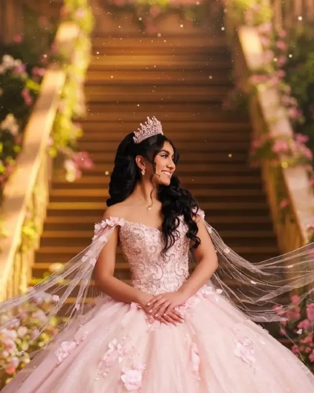 Quinceañera con corona y vestido rosado sentada en una escalera decorada con flores y luces cálidas.