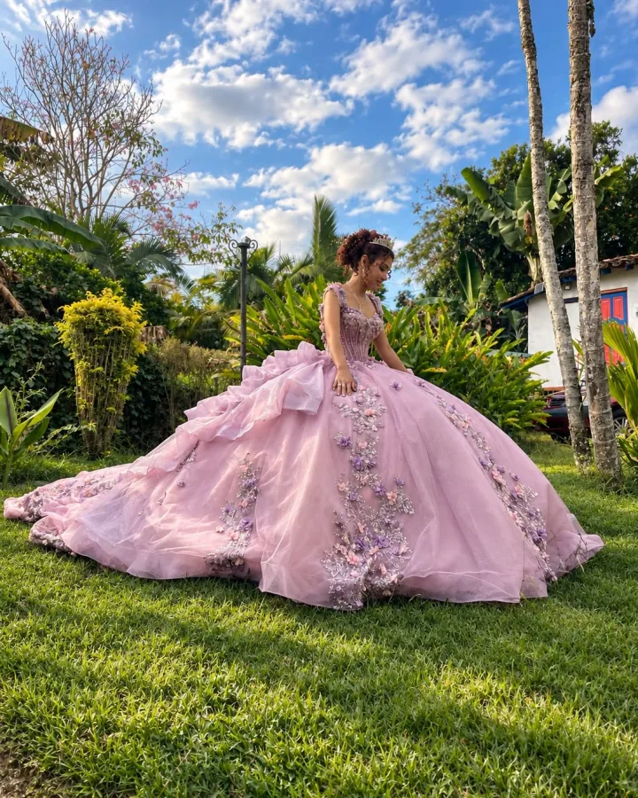 Quinceañera con peinado recogido y vestido rosa lila con flores aplicadas, sentada sobre el césped de un jardín.