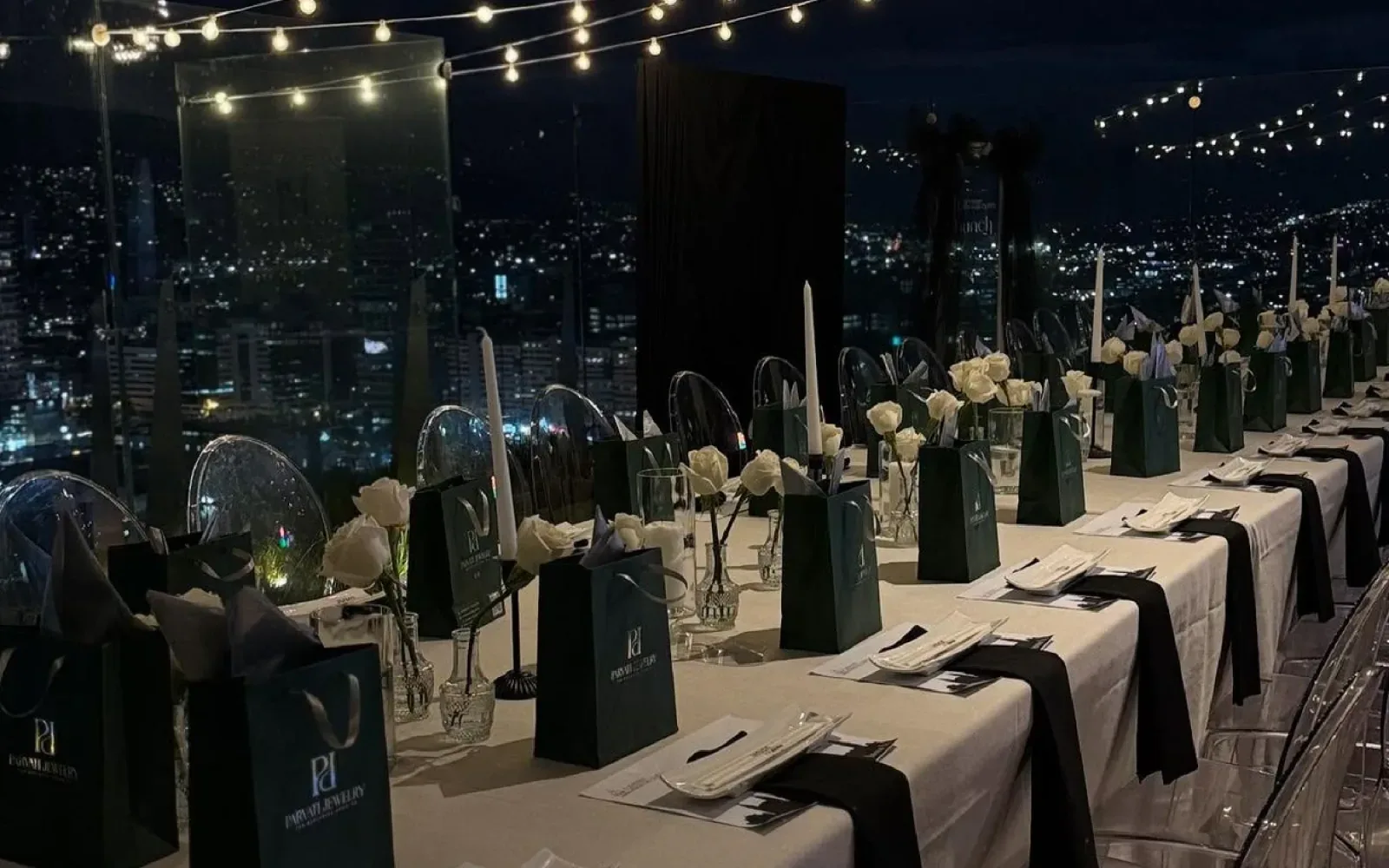 Mesa de evento con velas, flores blancas y luces colgantes, con una ciudad iluminada de noche al fondo.
