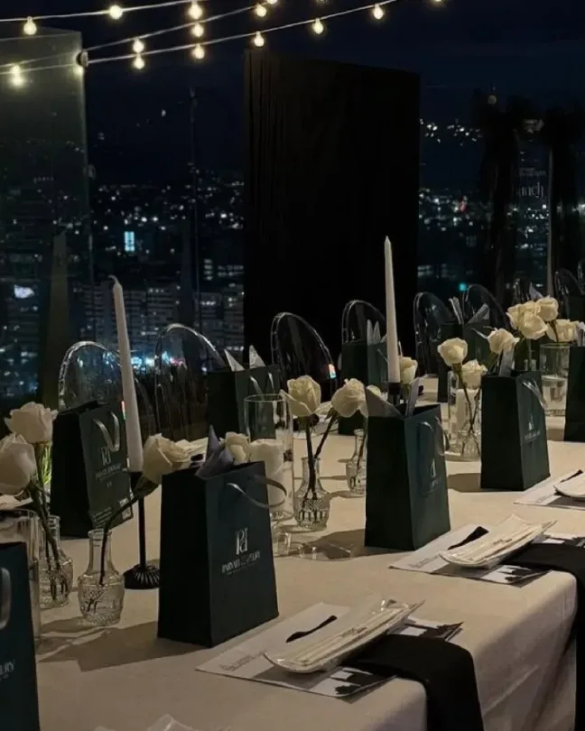 Mesa de evento con velas, flores blancas y luces colgantes, con una ciudad iluminada de noche al fondo.