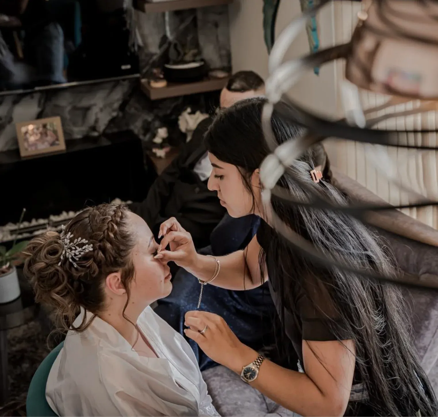 Maquilladora aplicando maquillaje de ojos a una novia con recogido trenzado, tocado plateado y bata blanca.