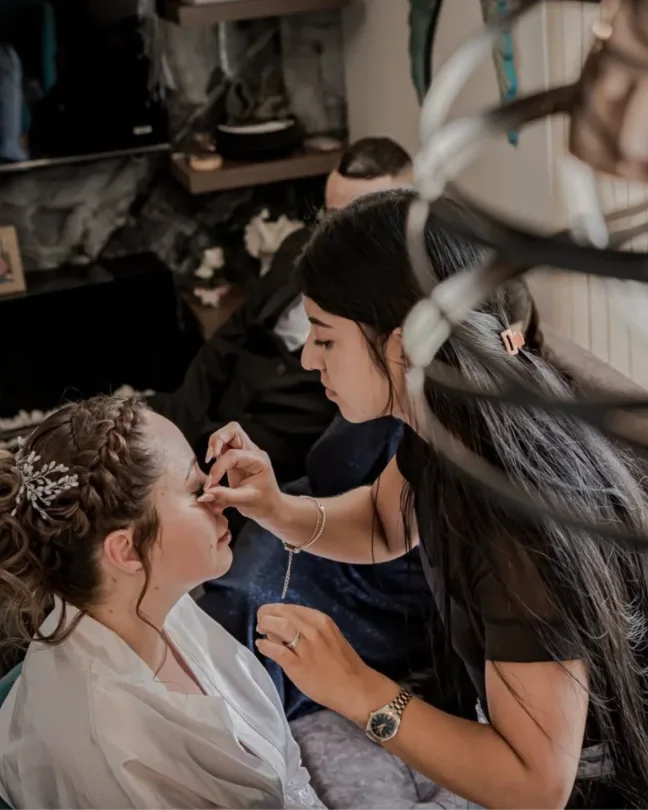 Maquilladora aplicando maquillaje de ojos a una novia con recogido trenzado, tocado plateado y bata blanca.