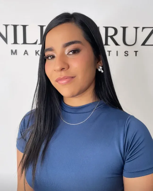 Maquillaje social suave en tonos tierra, con brillo en el lagrimal y labios nude rosados; cabello negro largo y liso y blusa azul, frente al letrero del estudio NilleCruz.