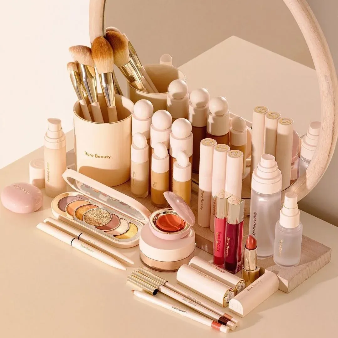 Brochas, sombras, labiales y otros cosméticos organizados frente a un espejo redondo.