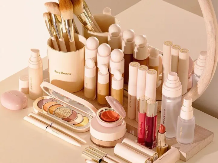 Brochas, sombras, labiales y otros cosméticos organizados frente a un espejo redondo.