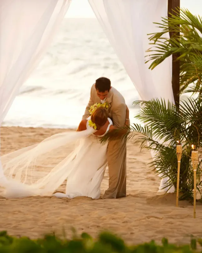 Pareja de novios abrazándose en la playa bajo un arco con cortinas blancas y flores.