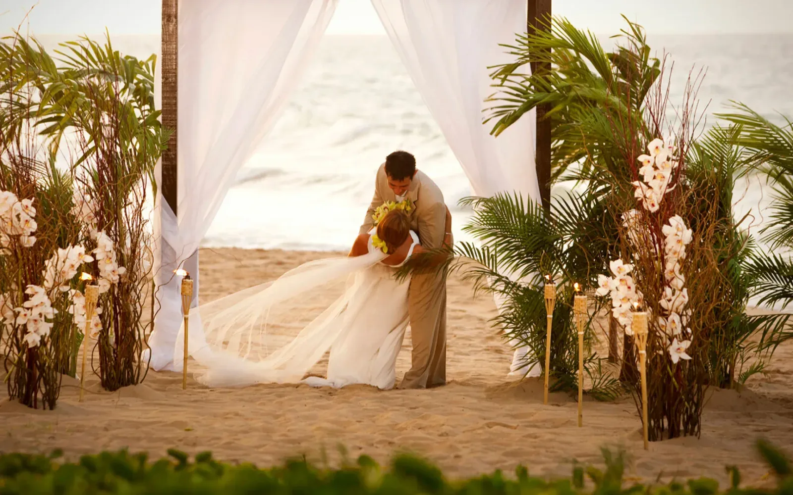 Pareja de novios abrazándose en la playa bajo un arco con cortinas blancas y flores.